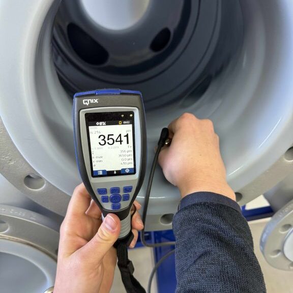 Hand hält ein QNix Schichtdickenmessgerät mit externer Sonde bei einer Messung an einem grauen, industriellen Bauteil (Messwert 3541 µm)