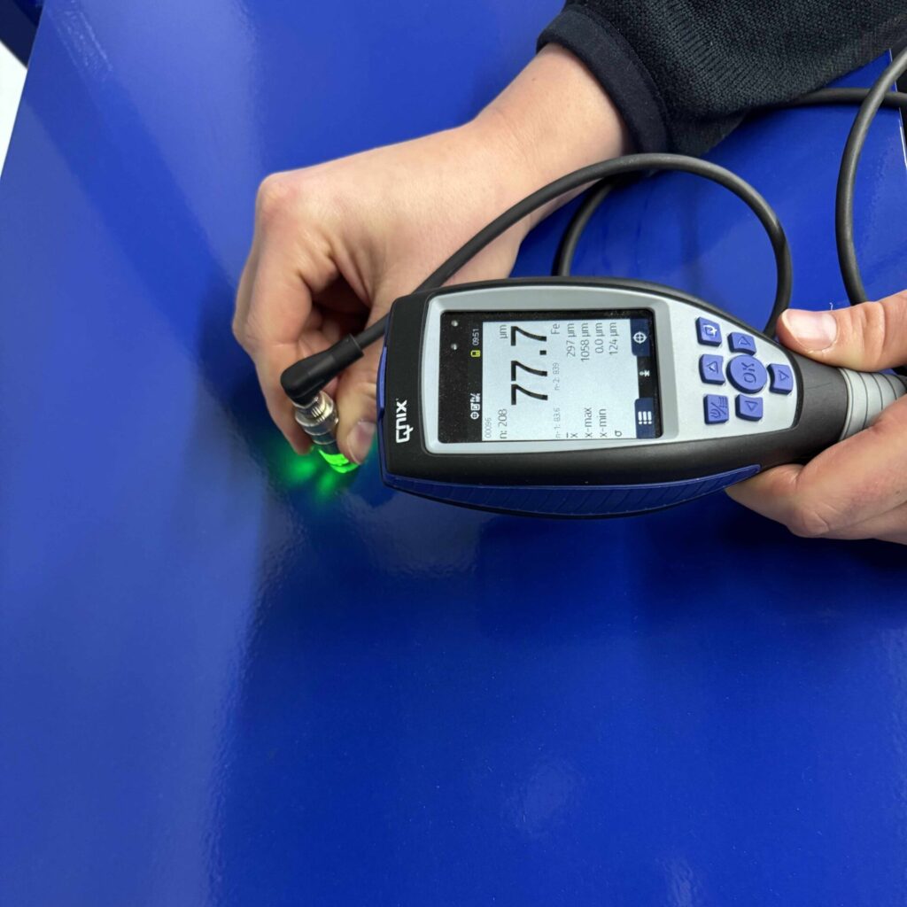 Hand hält ein QNix Schichtdickenmessgerät mit externer Sonde bei einer Messung auf einer blauen Oberfläche (Messwert 77,7 µm)