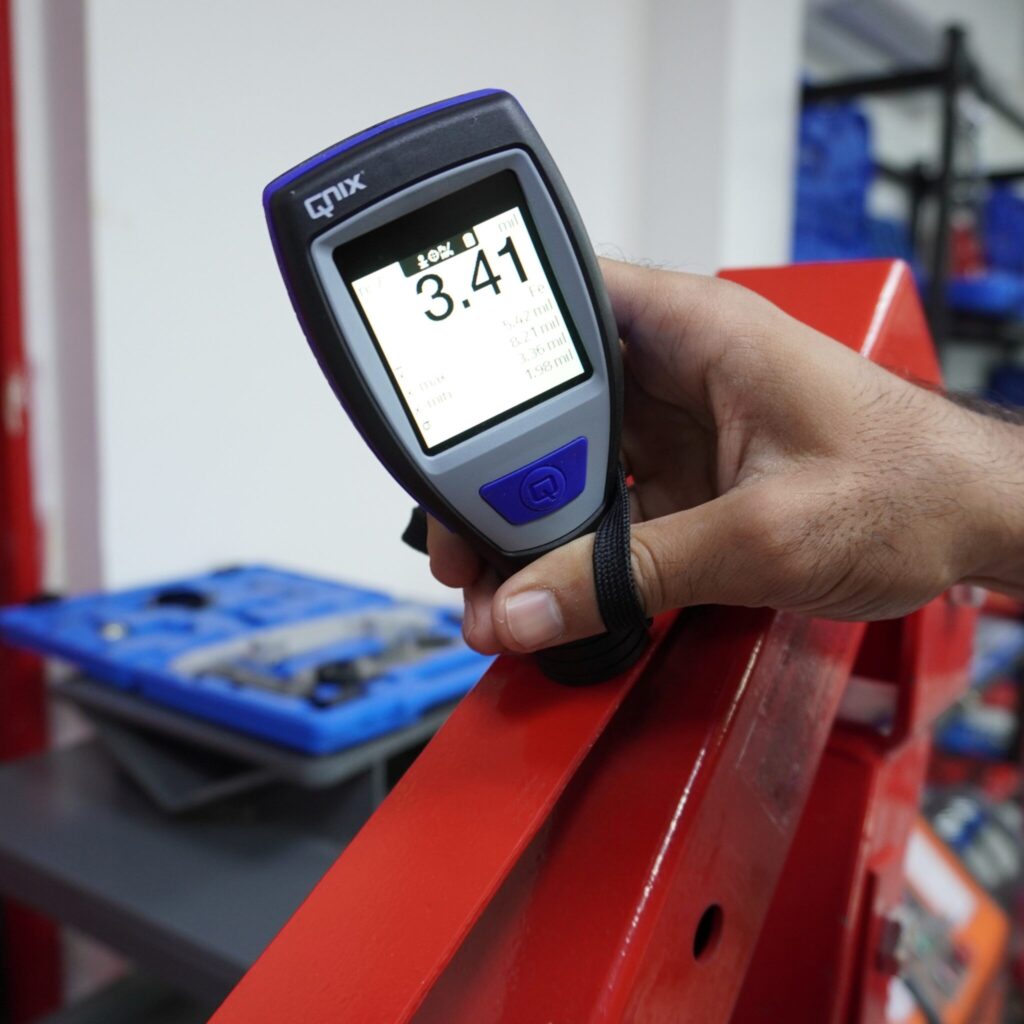 Hand hält ein QNix Schichtdickenmessgerät bei einer Messung an einer roten Metallstruktur (Messwert 3,41 mil)