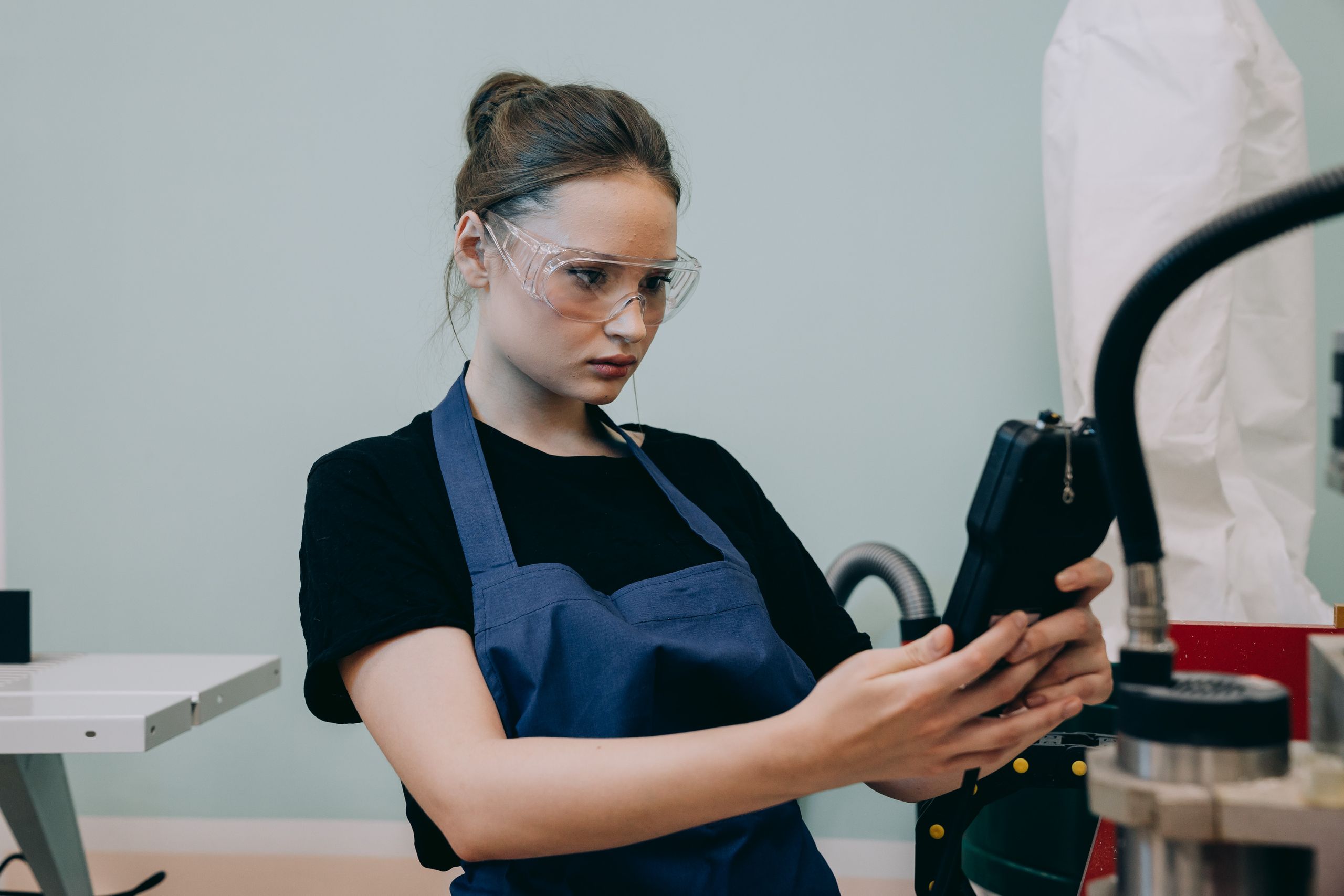 A woman in a blue apron uses a device in a workshop, wearing protective goggles.